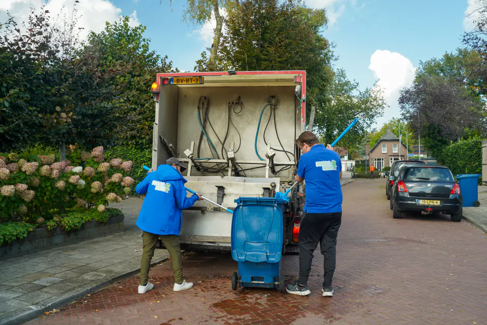 Twee medewerkers van Frisse Kliko reinigen samen een container bij de reinigingswagen in een woonstraat.